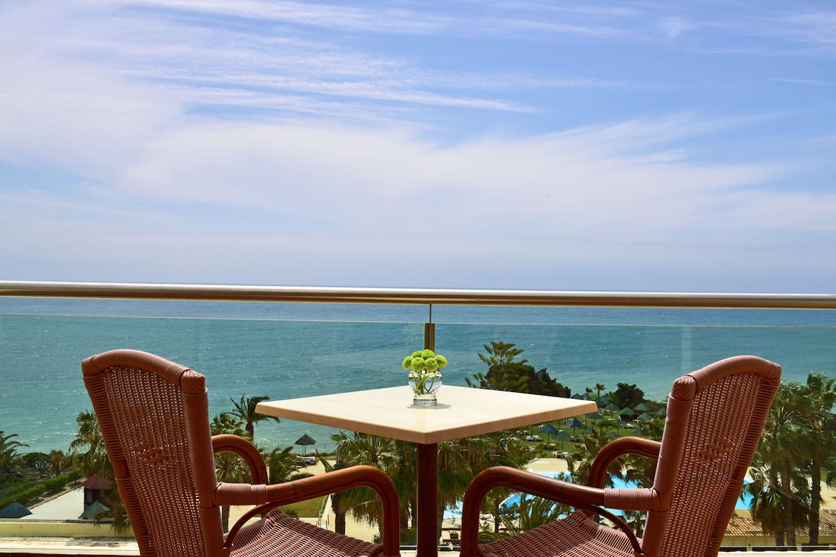 A balcony with wicker chairs and a table, overlooking a beach and the ocean.