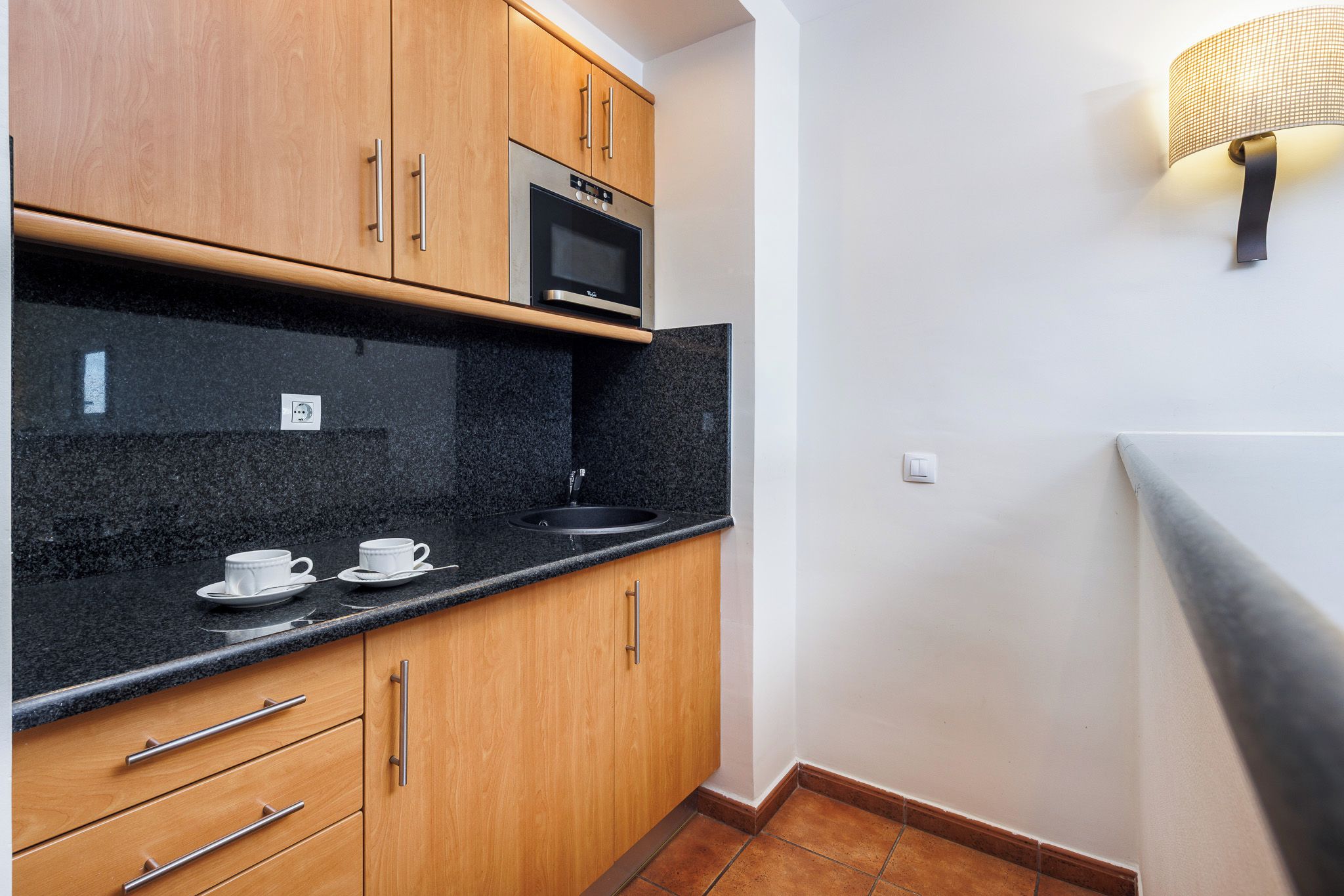 A kitchen with wooden cabinets, a black countertop, a microwave, and a wall-mounted light fixture.