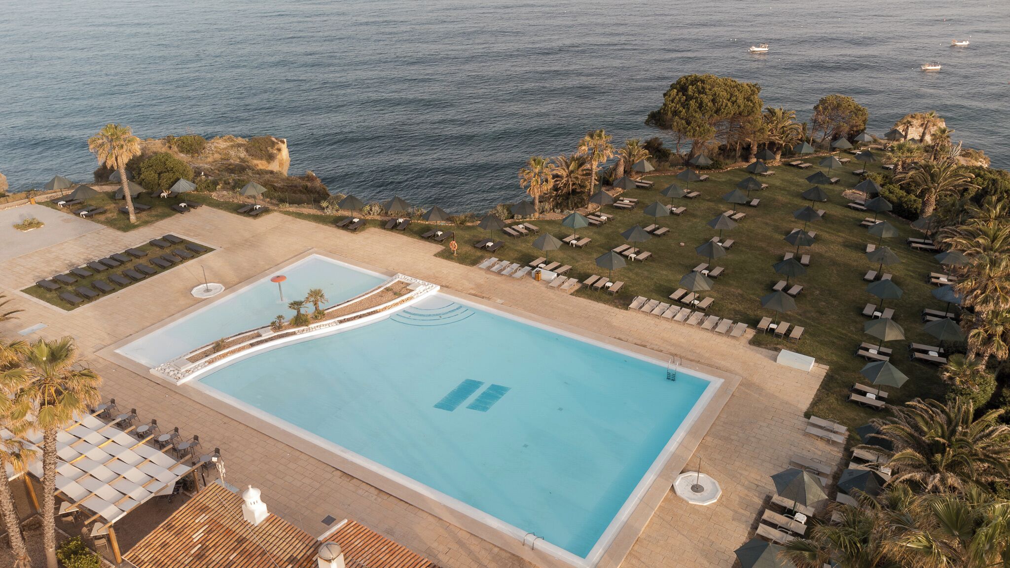 An aerial view of a resort with a large swimming pool, sun loungers, and umbrellas, surrounded by palm trees and overlooking the sea.