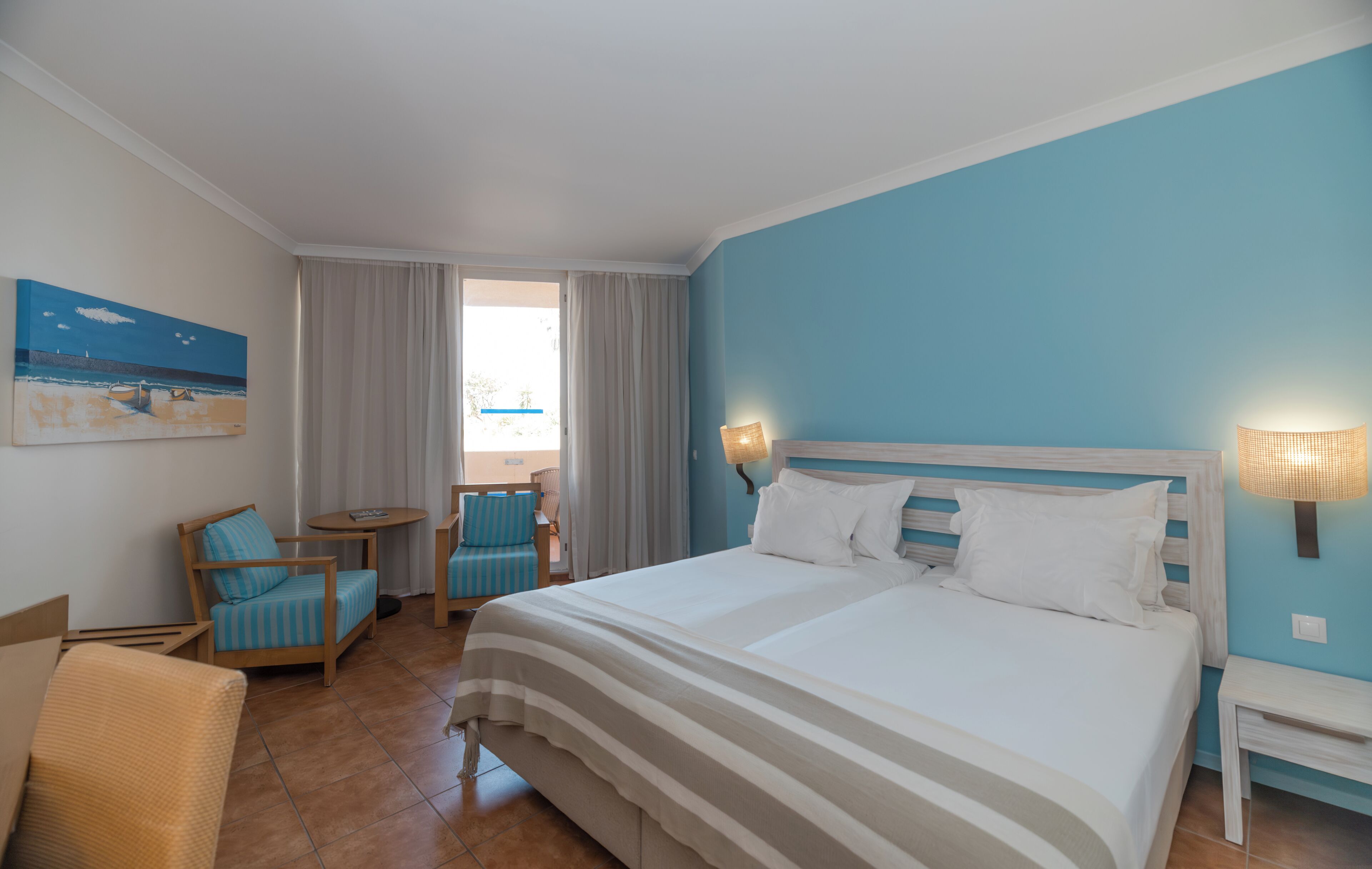 A hotel room with a bed, a chair, a small table, and a painting on the wall.