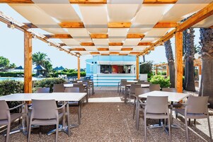 An outdoor cafe with tables and chairs, a bar in the background, and palm trees.