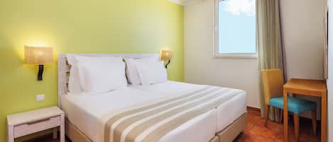 Habitación de hotel con cama, lámparas de noche, una mesita, una silla y un ventanal con cortinas.