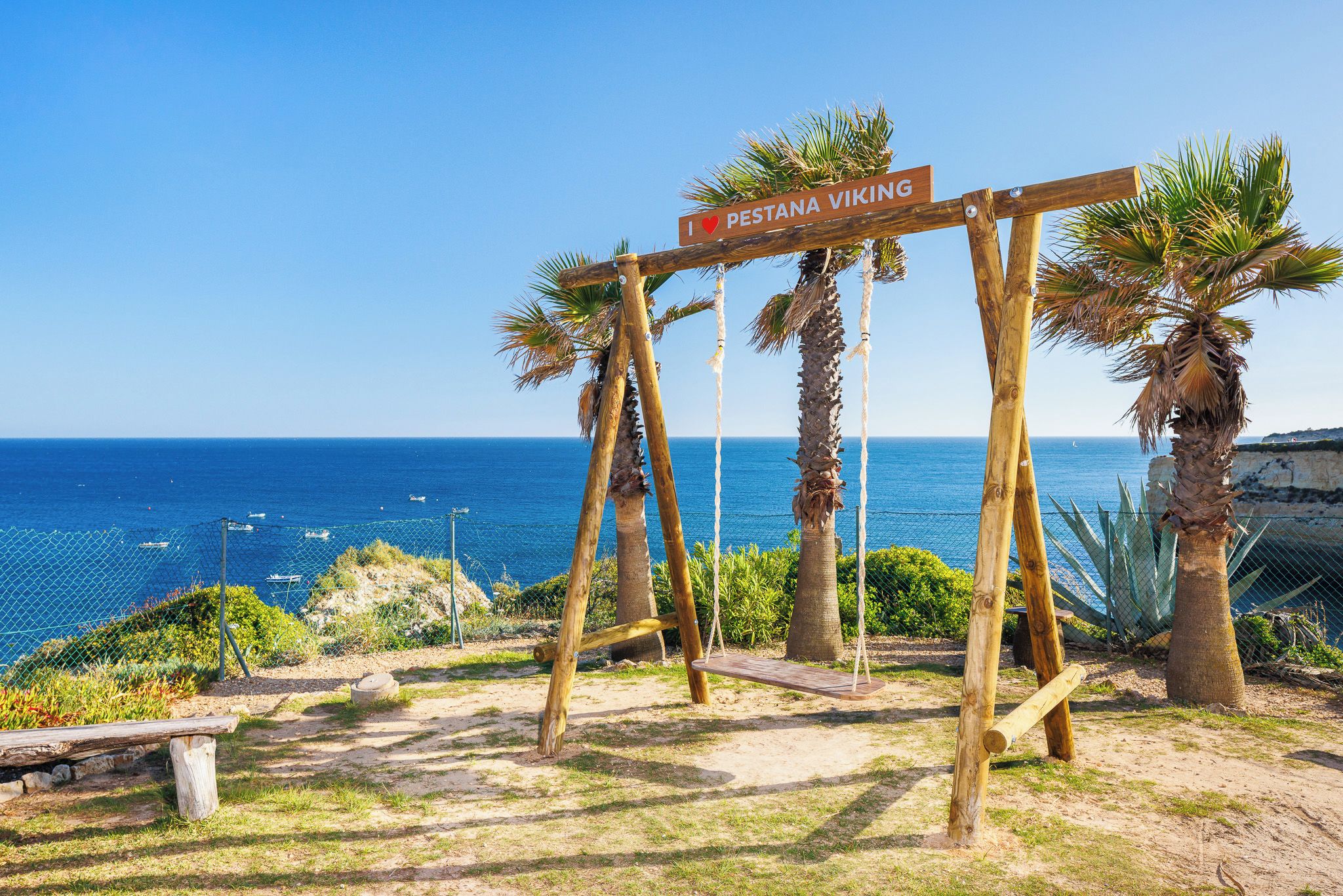 Un letrero de madera con la inscripción 'I ♥ PESTANA VIKING', con el mar y palmeras de fondo.