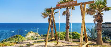 Un letrero de madera con la inscripción 'I ♥ PESTANA VIKING', con el mar y palmeras de fondo.
