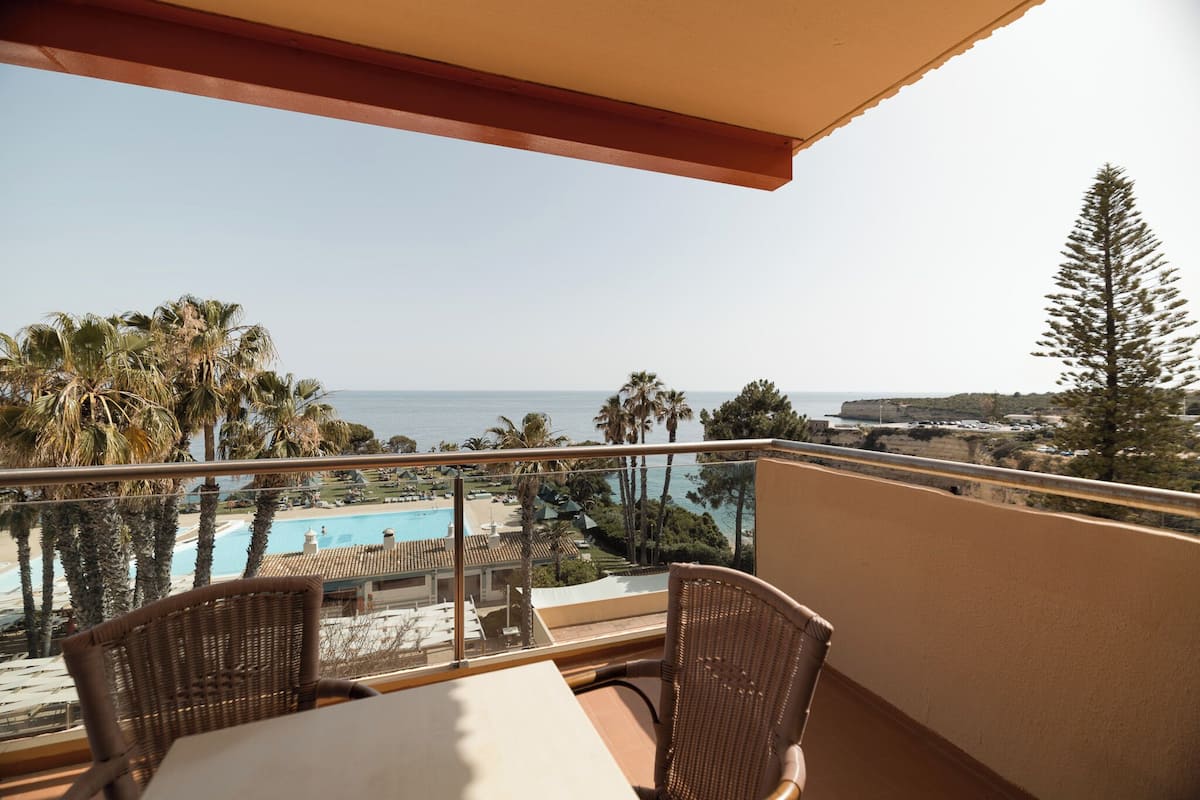 A balcony with wicker chairs and a table overlooking a pool and beach.