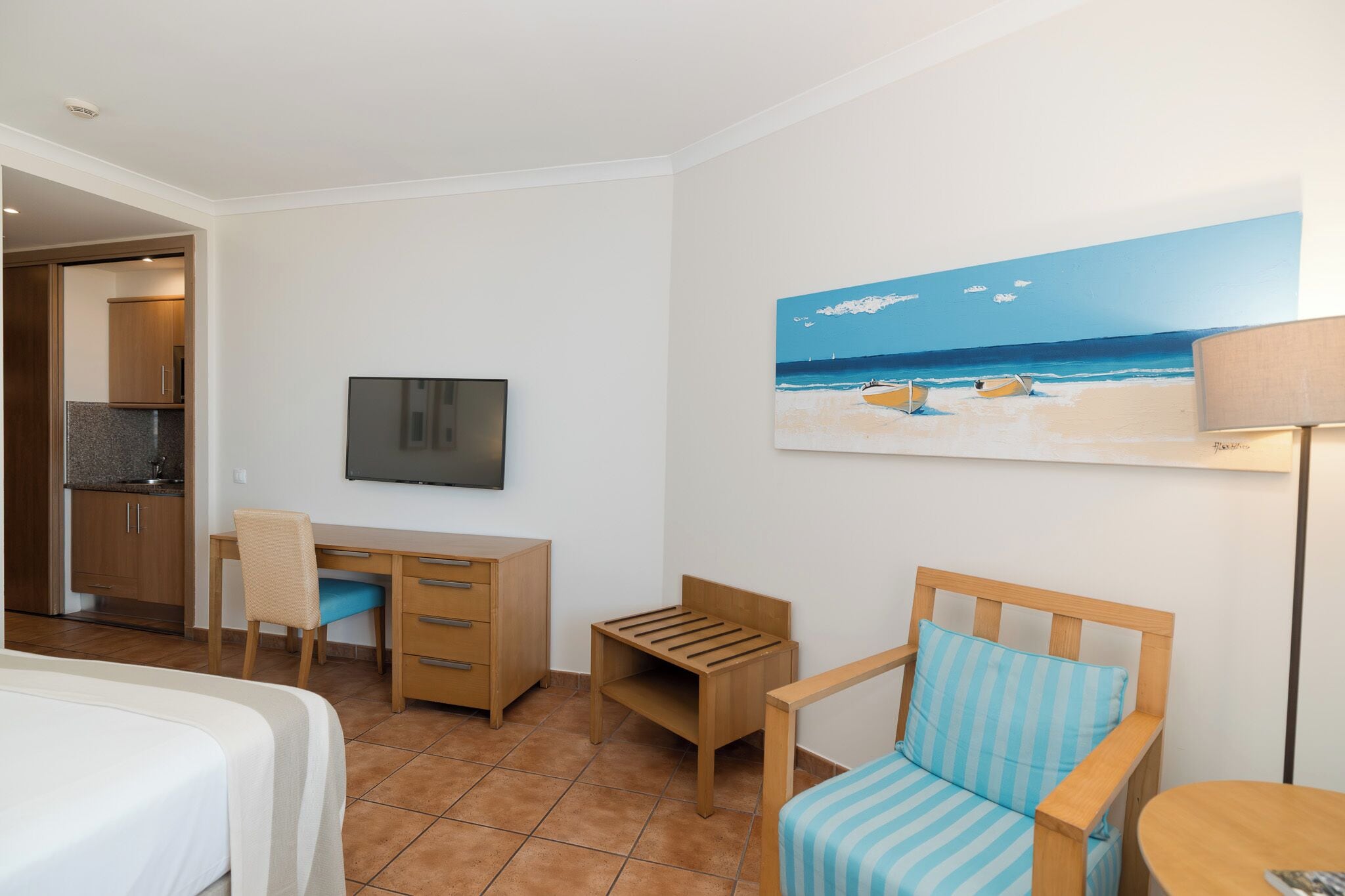 Une chambre d’hôtel comprenant un lit, un bureau, une chaise, une petite table et un tableau représentant une scène de plage.