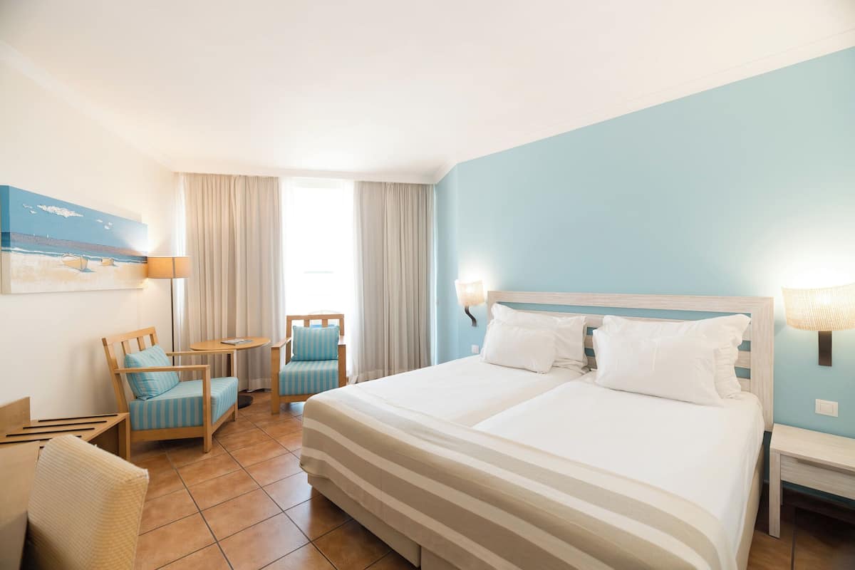 A hotel room with a large bed, two chairs, a small table, and a painting on the wall.