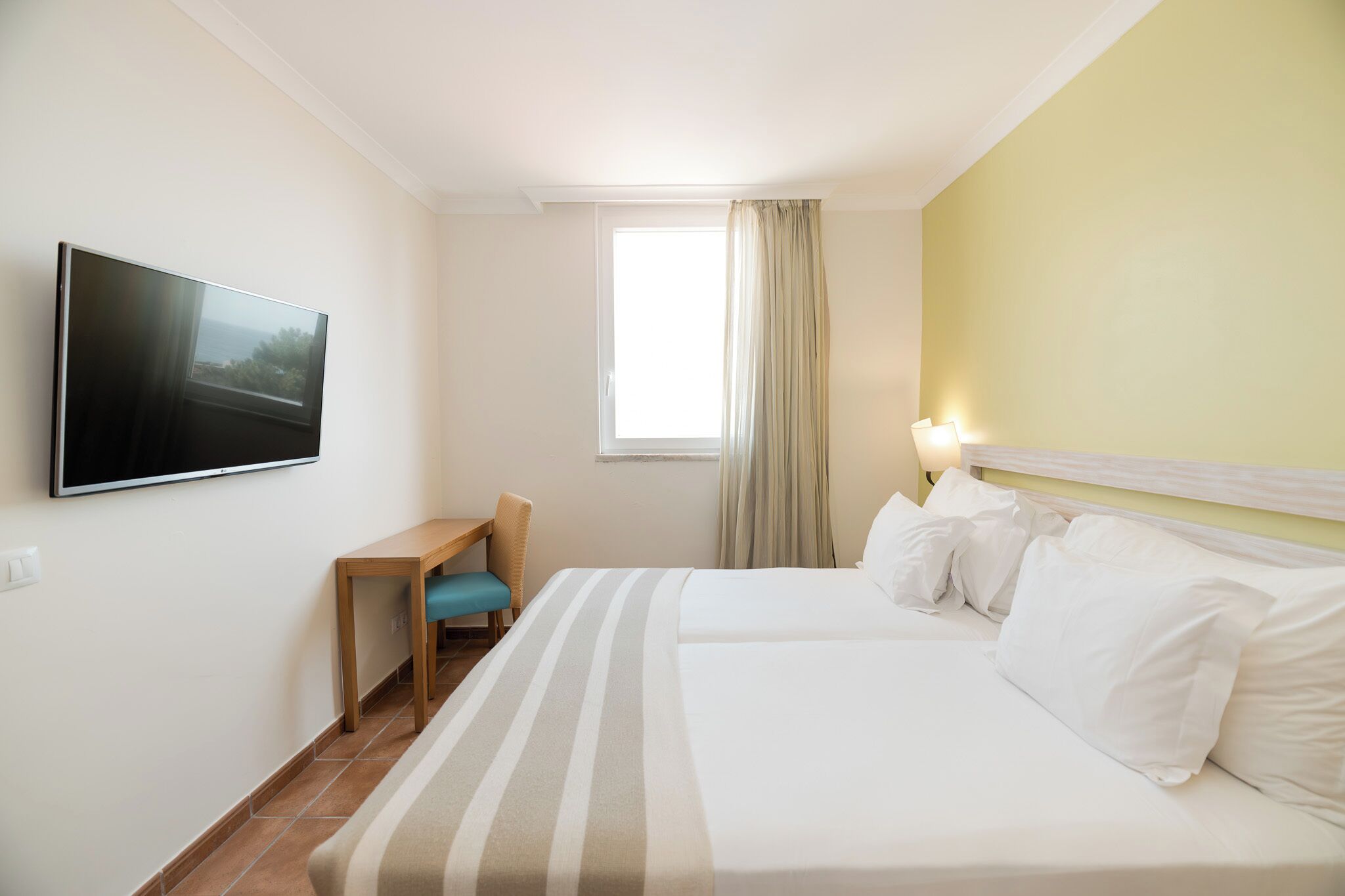 Habitación de hotel con cama, escritorio, silla, televisor montado en la pared y ventana con cortinas.