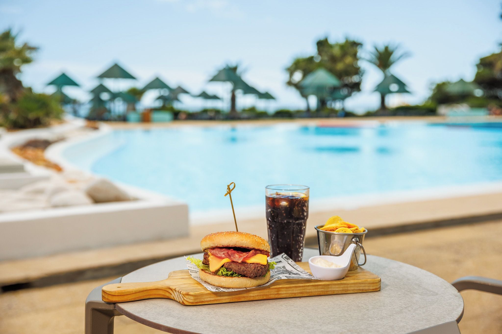 A meal with a burger, a drink, and chips by a poolside.