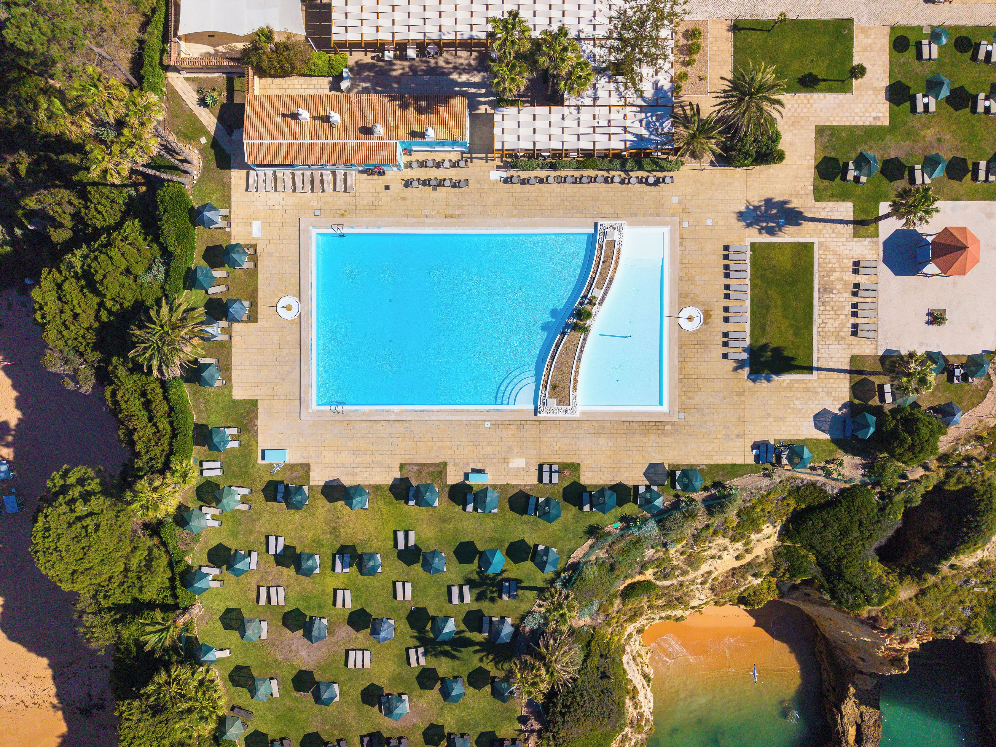An aerial view of a resort with a central swimming pool, multiple lounge areas with umbrellas, and a beach area.