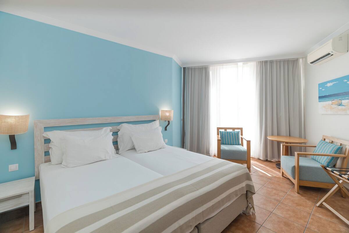 A hotel room with a large bed, a desk, a chair, and a view of the beach through the window.