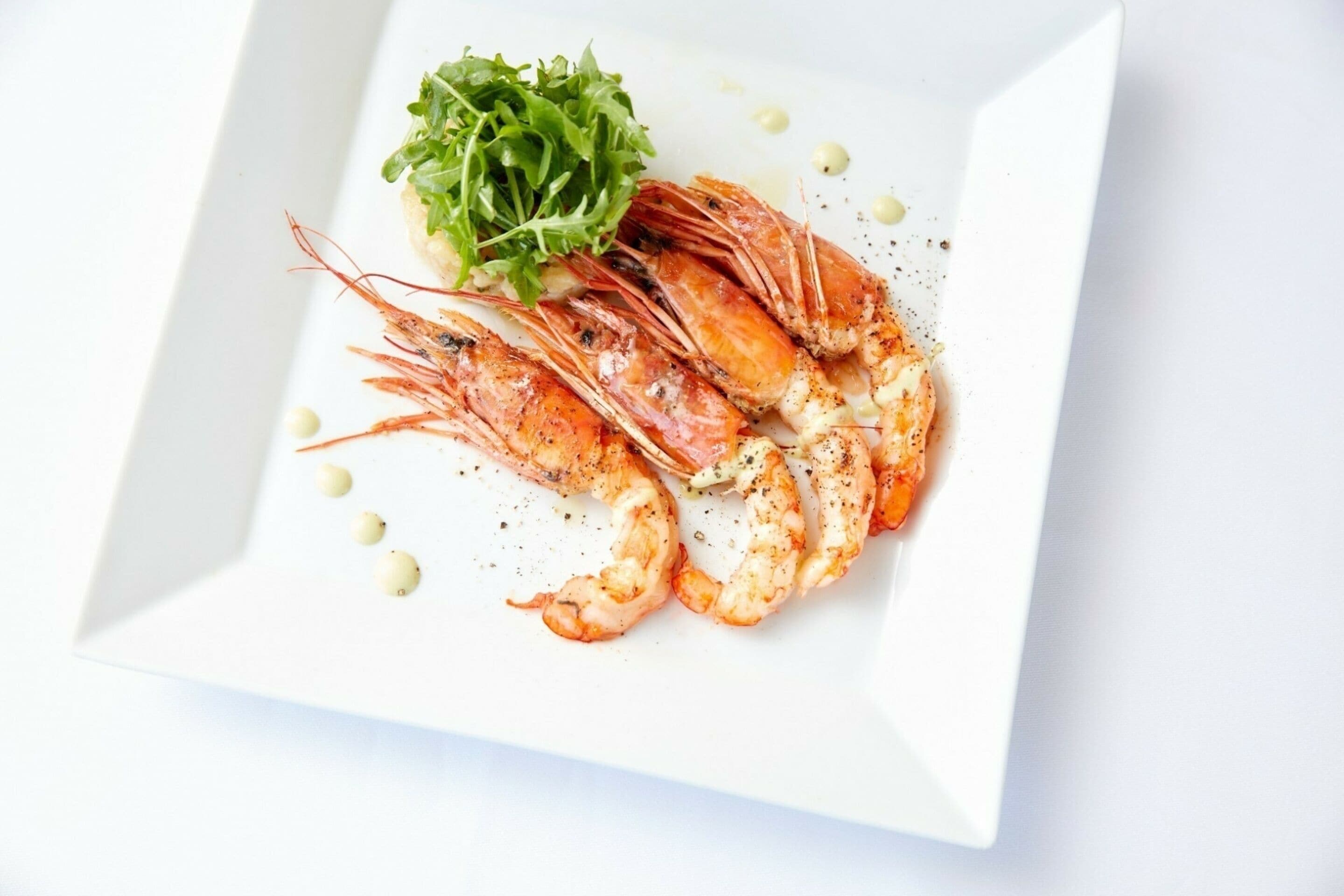 A plate of cooked shrimp with a garnish of greens and small round items, served on a white square plate.