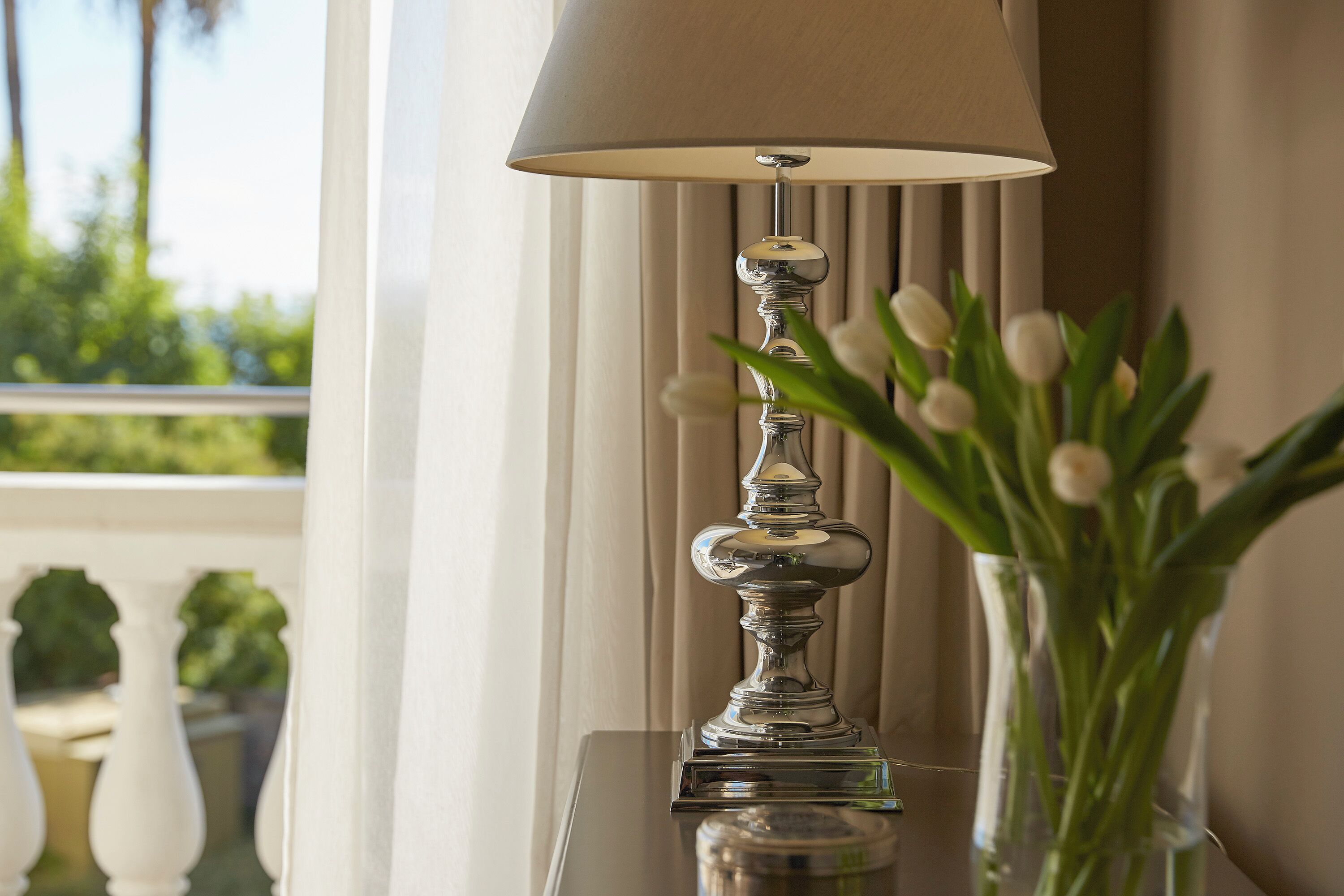 Eine Tischlampe mit beigem Schirm, einem klaren Glasfuß und einer Vase mit weißen Blumen.