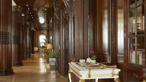Un elegante corridoio con imponenti colonne in legno intagliato, un tavolo con piano in marmo e un telescopio in ottone, e una libreria piena di libri.