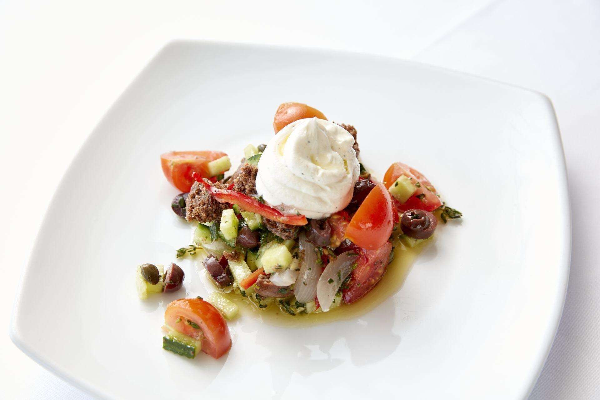 Un plato cuadrado blanco con una ensalada cubierta por una cucharada de salsa blanca, posiblemente yogur o crema agria, y adornada con rodajas de tomate y pepino.