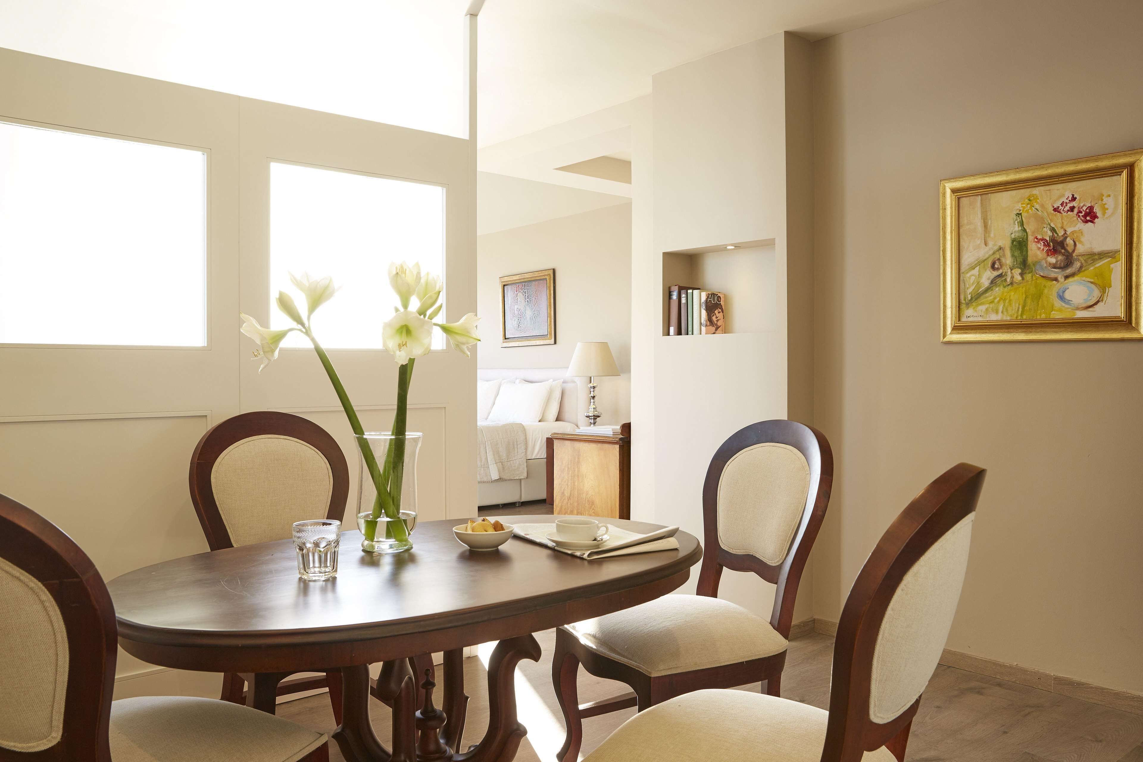 A dining area with a round table, four chairs, a vase with flowers, and a painting on the wall.