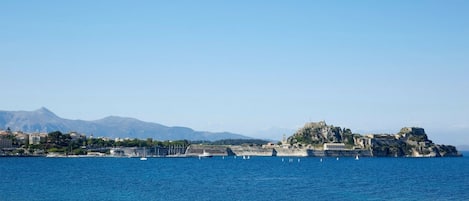 A coastal scene with boats on calm waters, a rocky island with buildings, and mountains in the background.