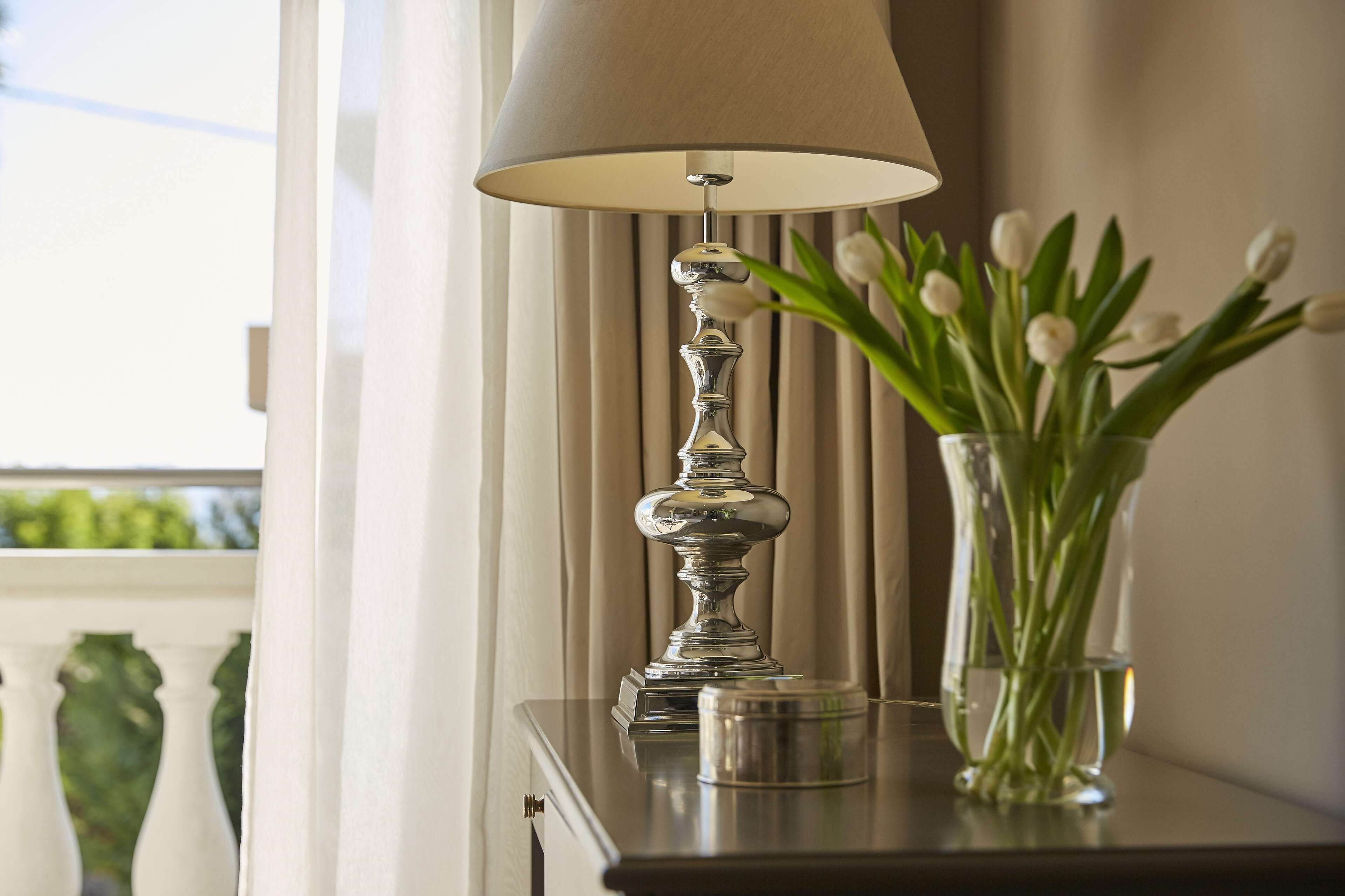 A lamp with a beige shade on a wooden table next to a vase with white flowers.