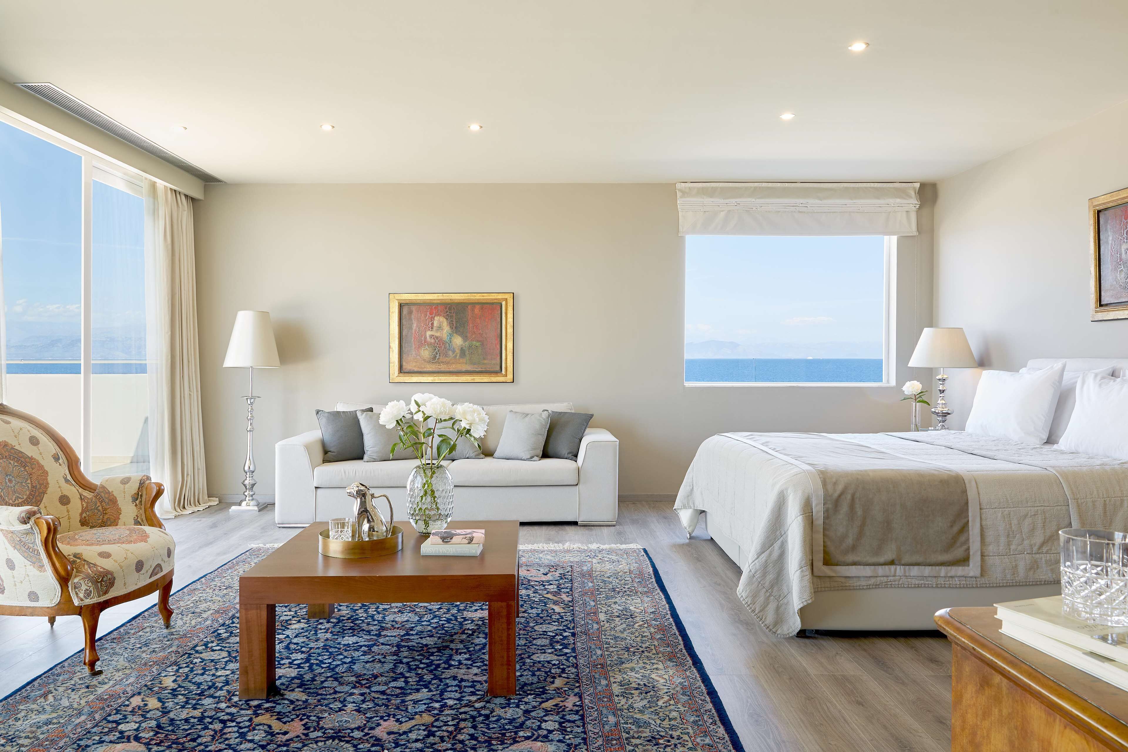 Une chambre d’hôtel avec une grande fenêtre, un canapé blanc, un lit, une table basse en bois et un tapis à motifs.
