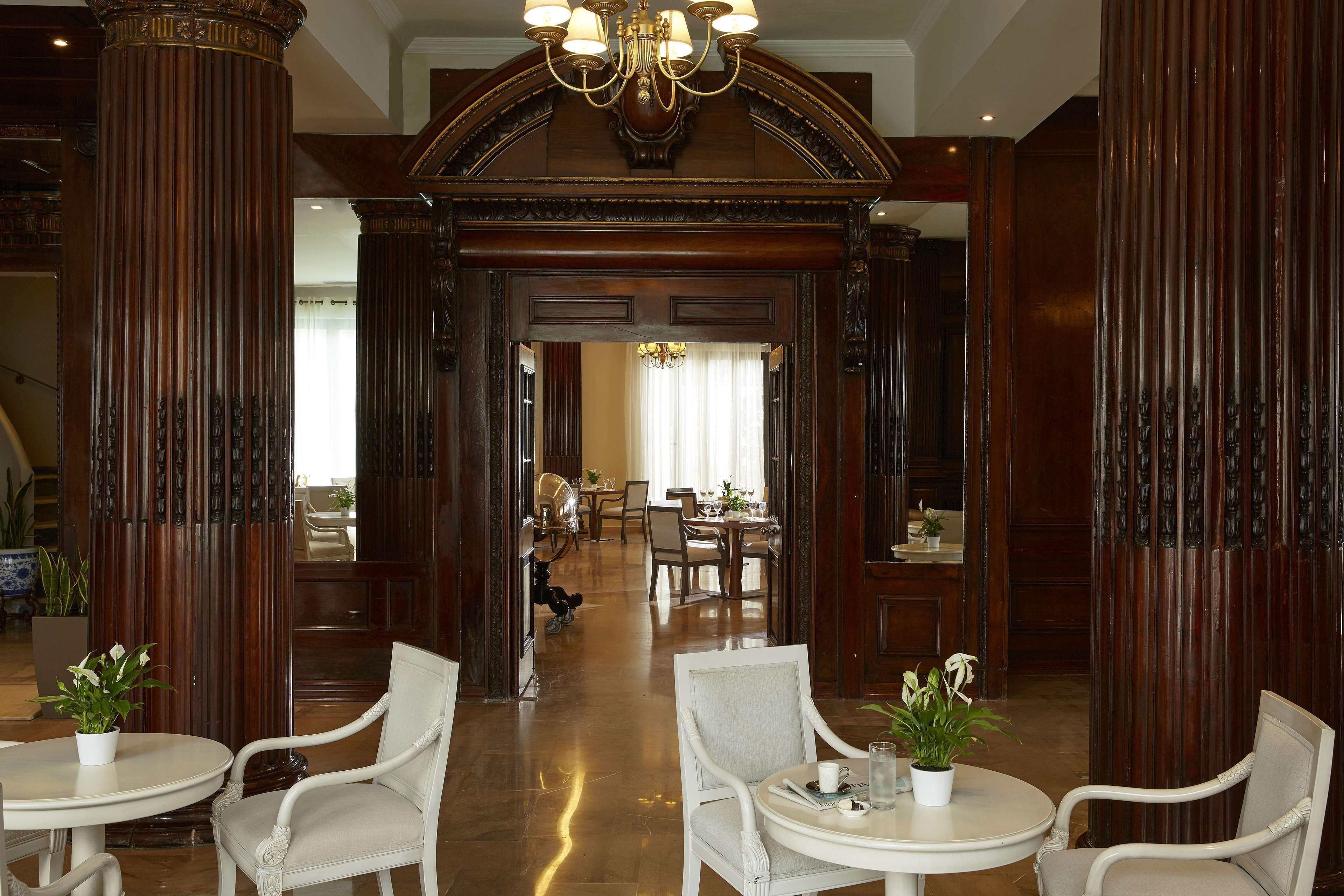 Eine großzügige Lobby mit hohen Holzsäulen, eleganten Kronleuchtern und weißen Polstersesseln um kleine Tische mit Topfpflanzen.