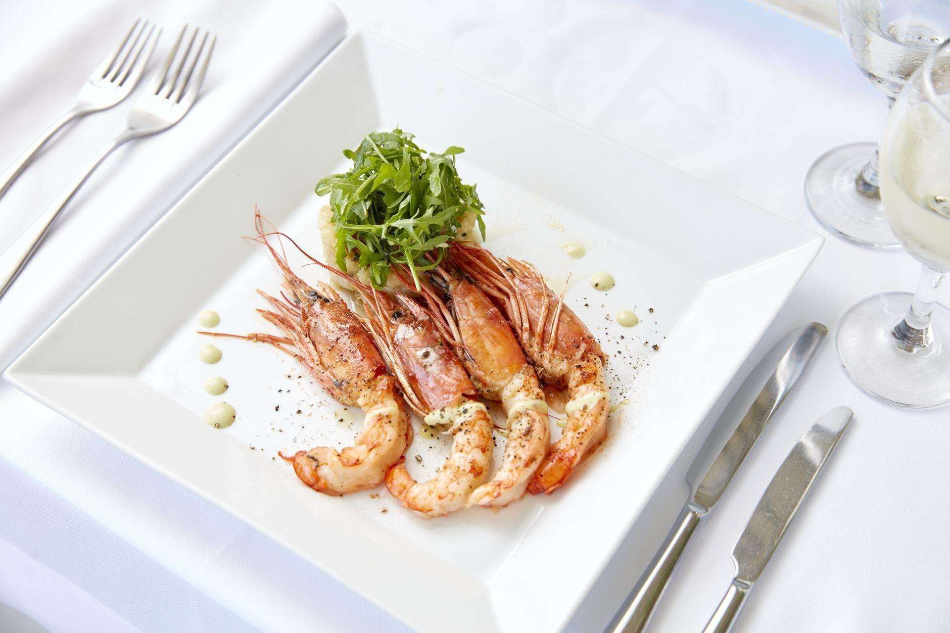 A white plate with cooked shrimp and greens, accompanied by silverware and a glass of white wine on a white tablecloth.