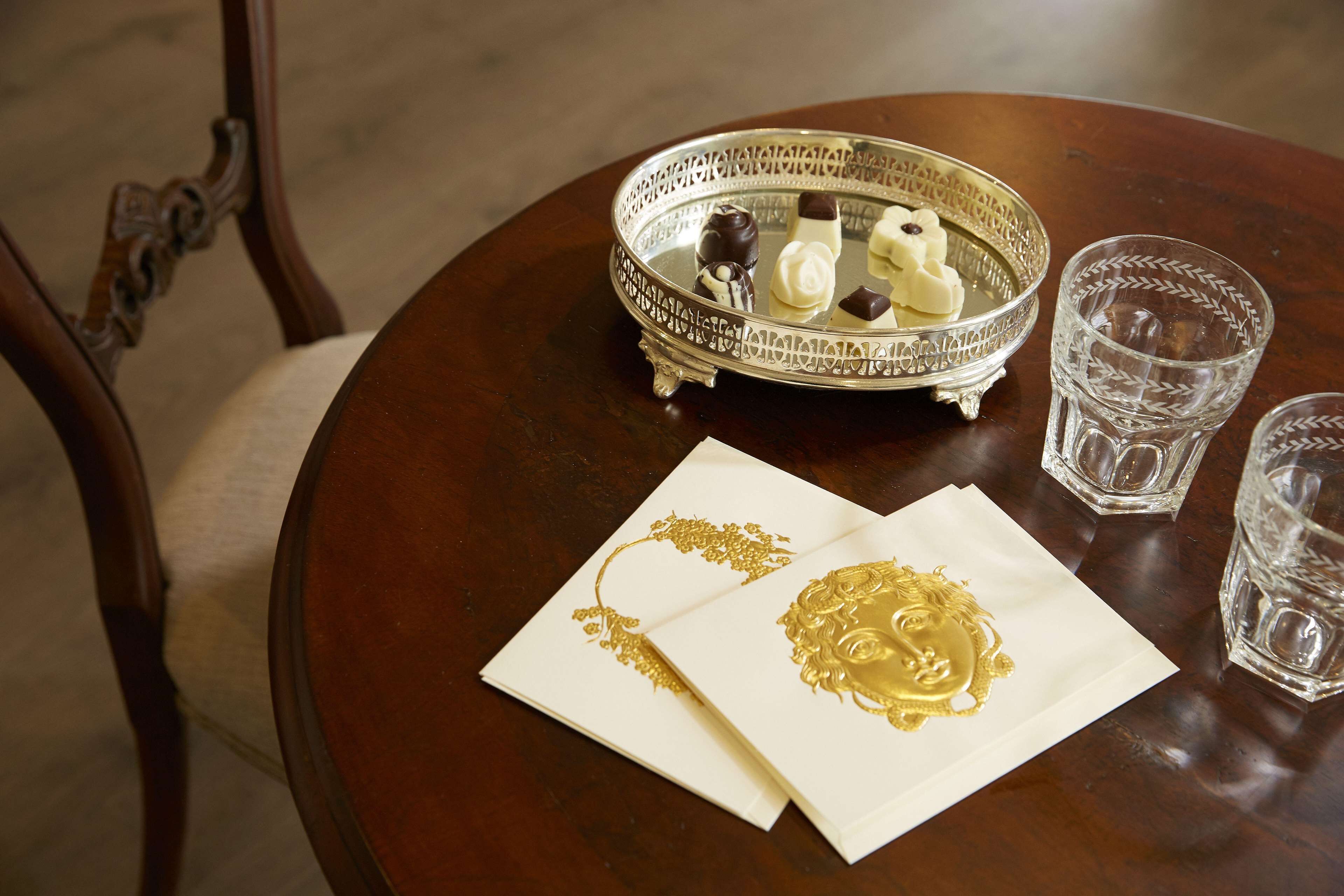 Uma mesa de madeira redonda com uma bandeja de prata decorativa, com chocolates e duas taças de vidro.