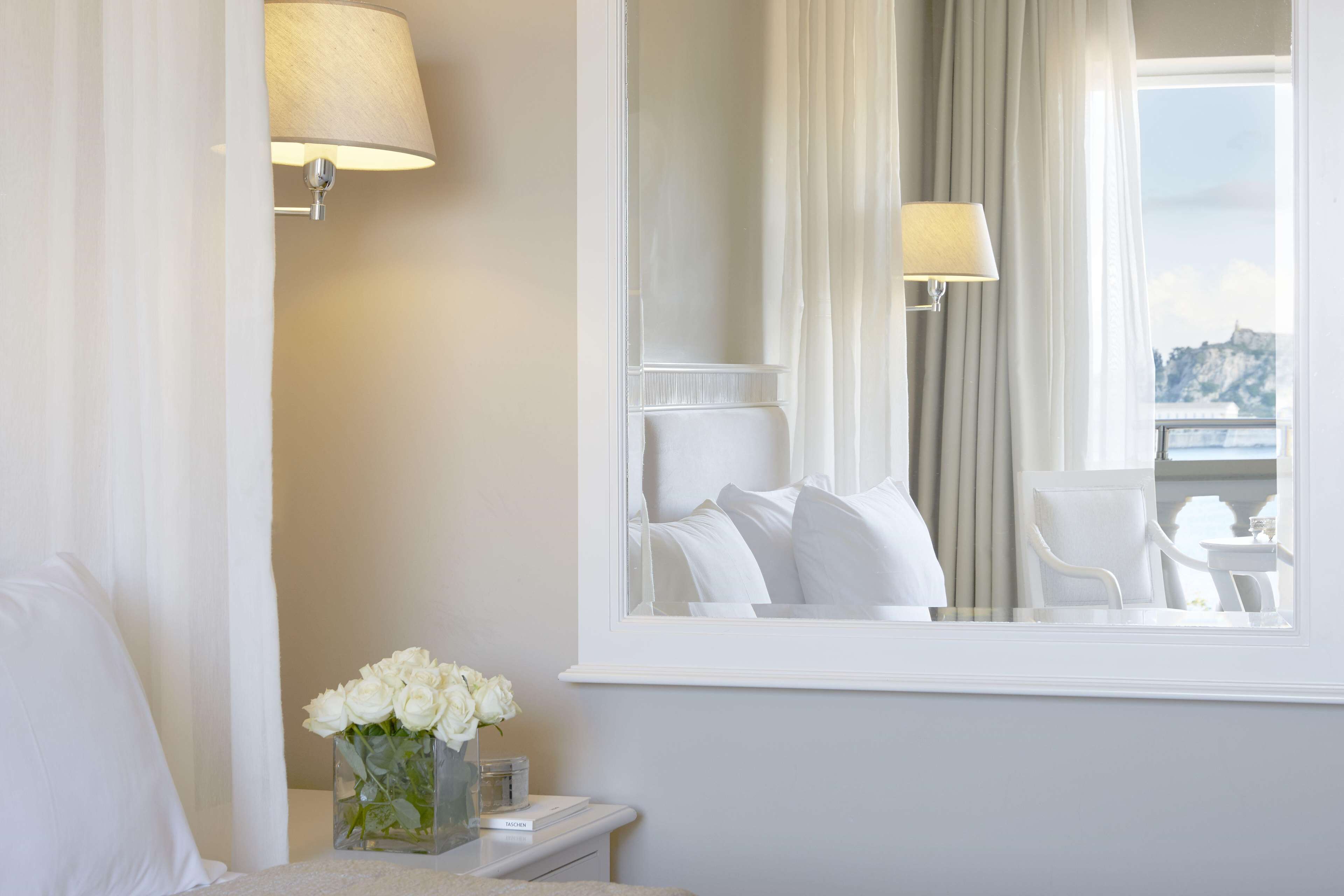 Habitación de hotel con cama, mesita de noche con un florero de flores blancas, ventana con vista a un cuerpo de agua y un edificio, y una lámpara en la pared.