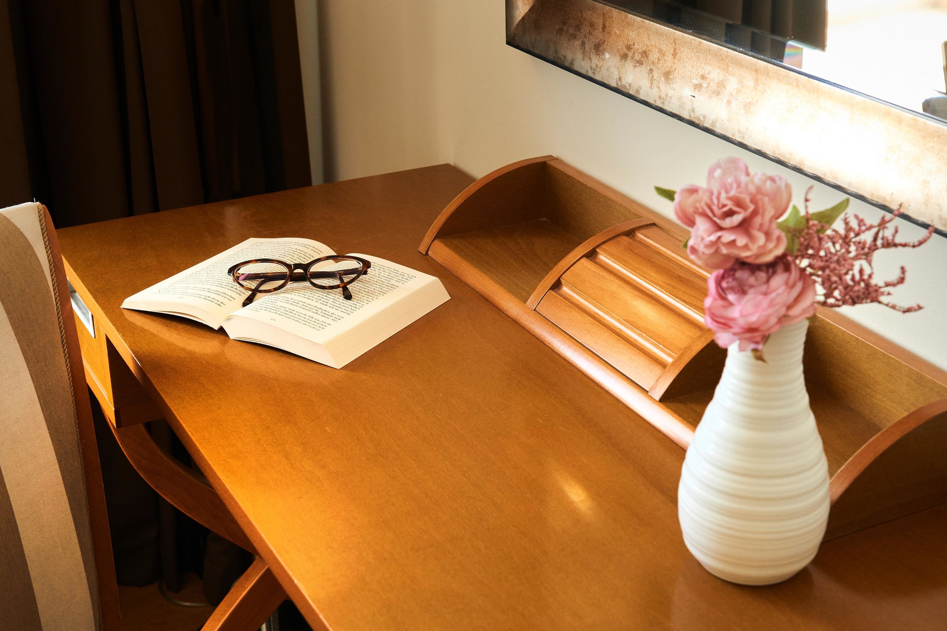 Un escritorio de madera con un libro, gafas y un florero con flores.