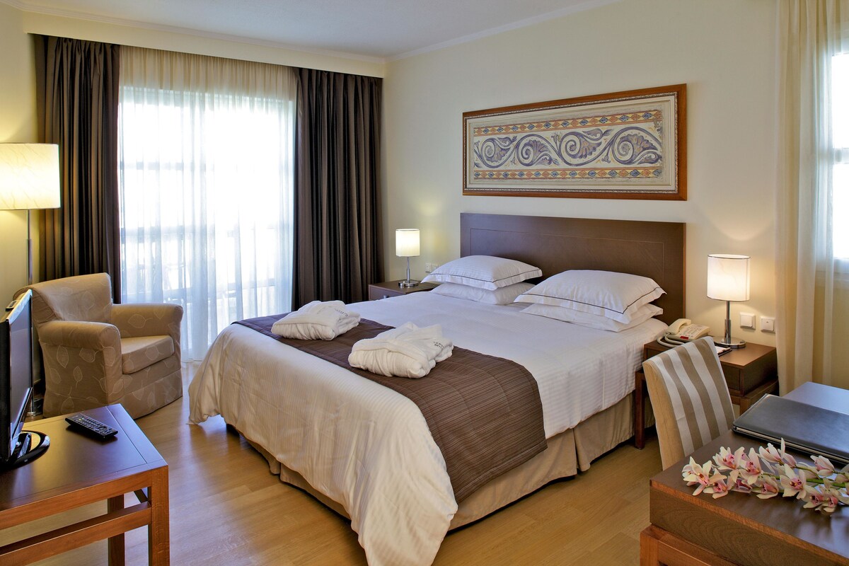 A hotel room with a large bed, two bedside tables, a chair, a desk, a telephone, and a decorative piece on the wall.