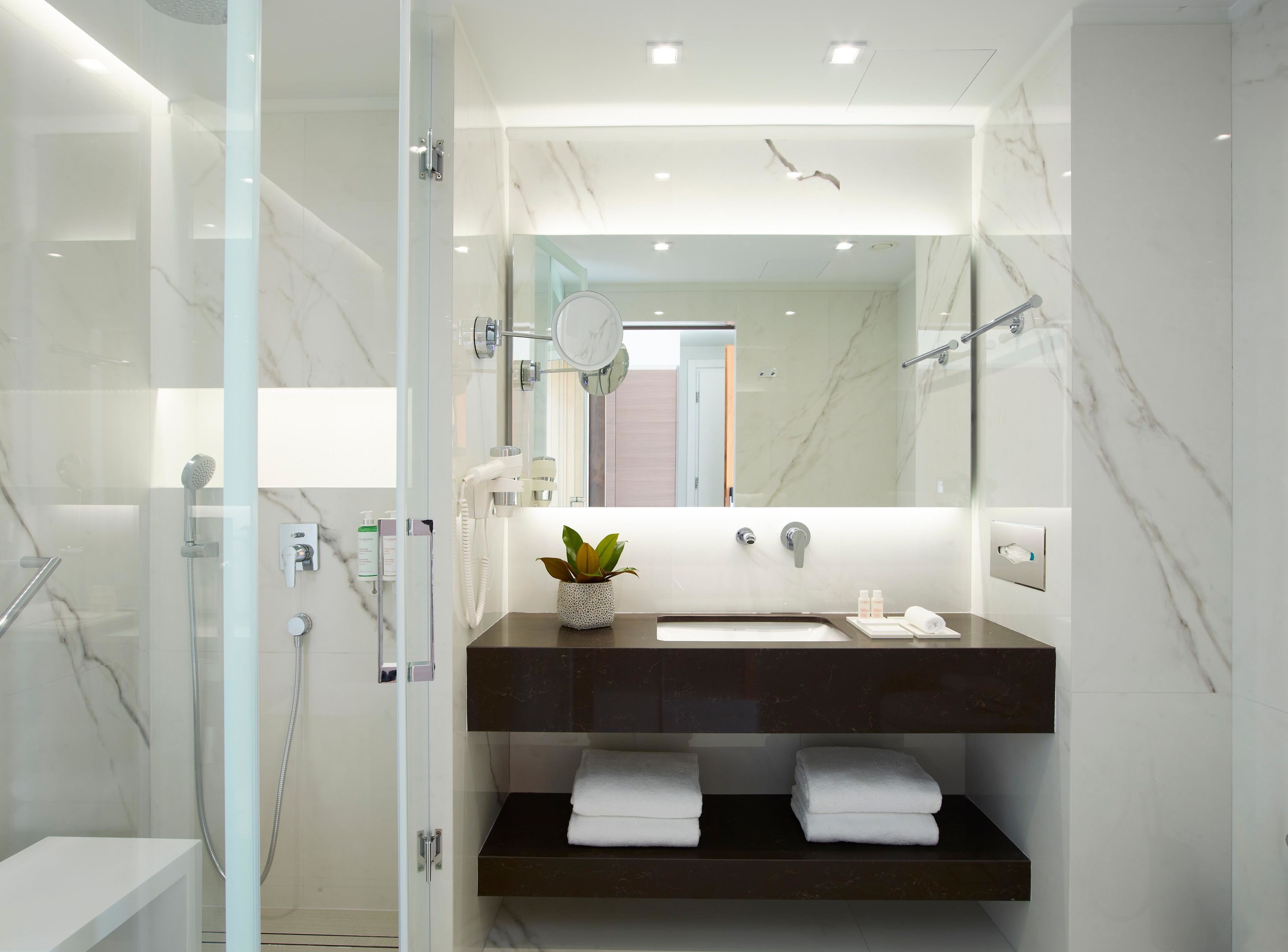 A modern bathroom with a glass-enclosed shower, a marble-tiled wall, a dark countertop sink, and a mirror above it.