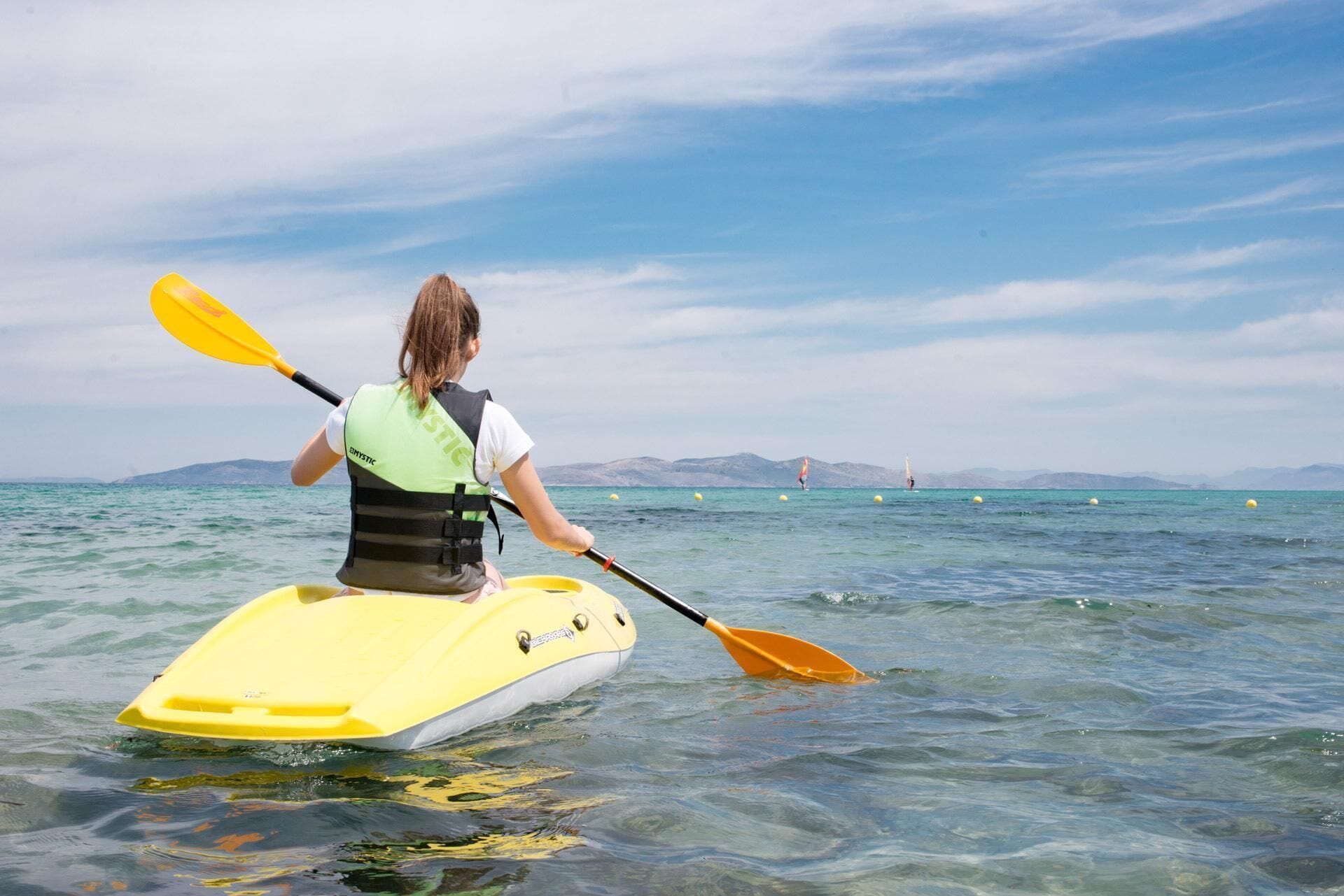 Une personne pratiquant le kayak par une journée ensoleillée, sur une eau calme, avec des montagnes qui se dessinent à l'horizon.