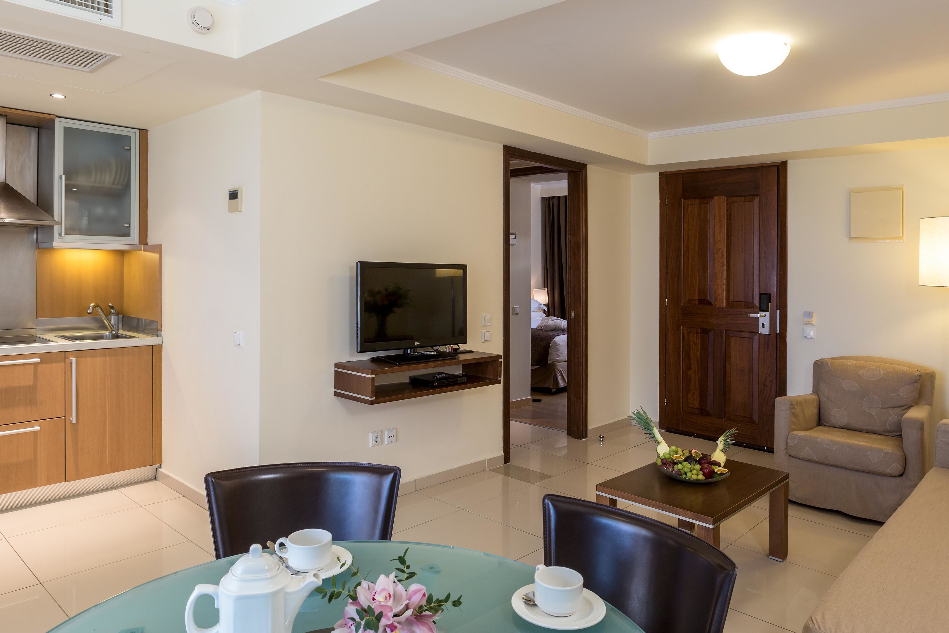A modern living room with a television, a dining table with chairs, a kitchen area with cabinets, and a wooden door.