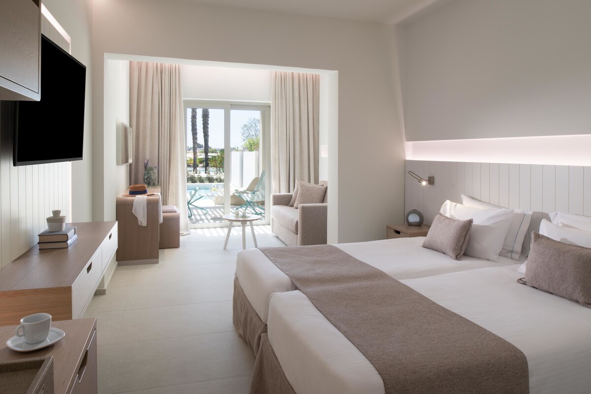 A hotel room with a bed, a desk with a cup and saucer, a chair, a TV, and a view of a pool and greenery.
