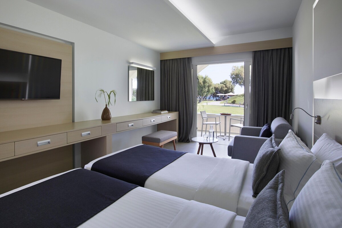 A hotel room with two beds, a desk, a TV, a mirror, and a view of the outdoors.