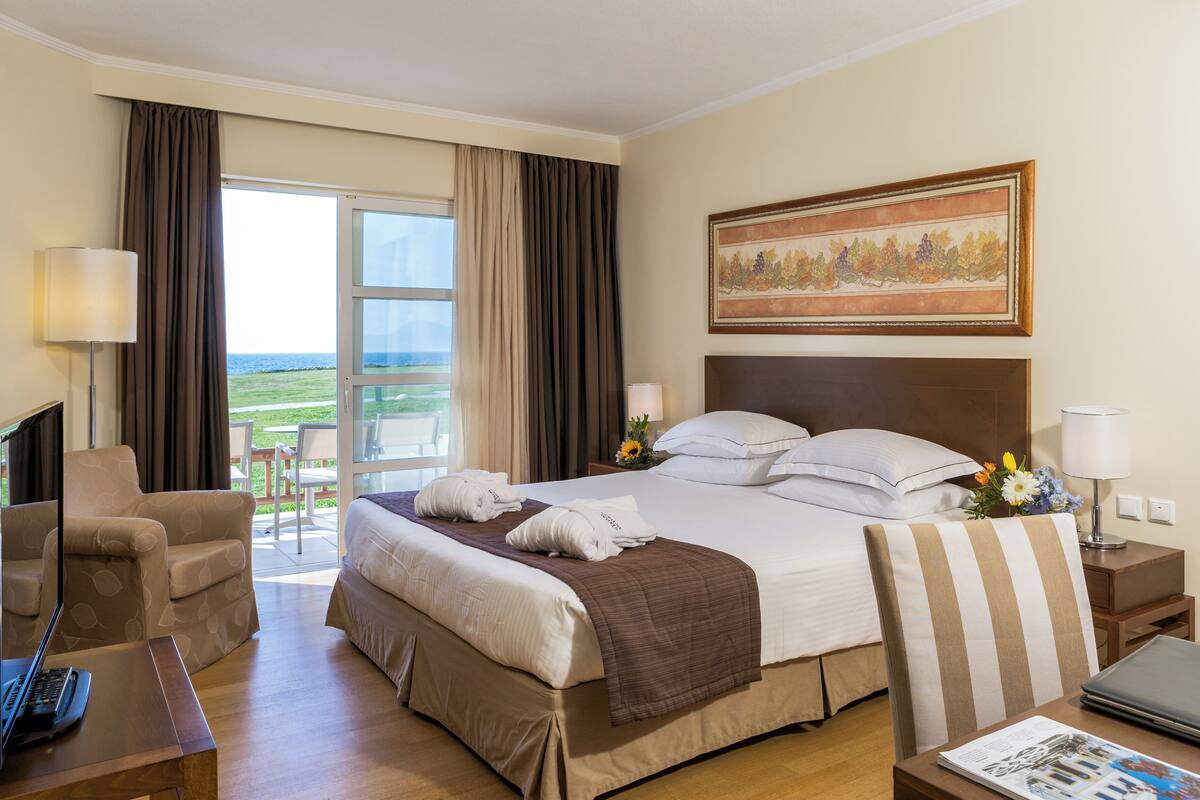 A hotel room with a bed, a desk, a chair, a TV, a window with curtains, a painting, and a view of the outdoors.