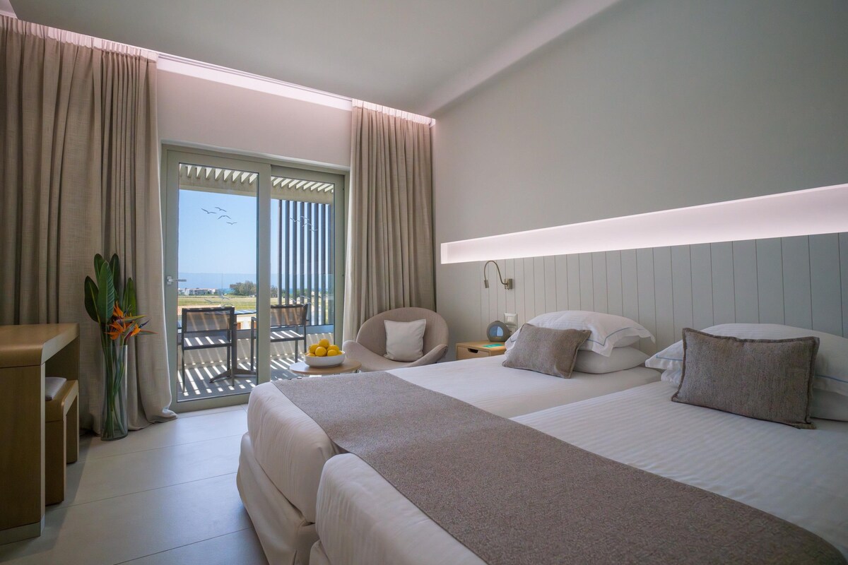 A hotel room with two beds, a desk, a chair, a nightstand, and a view of the outdoors.