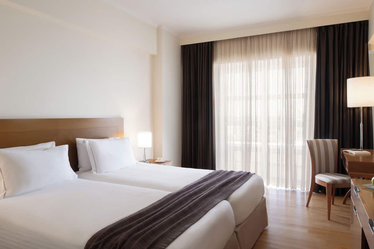 A hotel room with two beds, a desk with a chair, a lamp, and a large window with curtains.