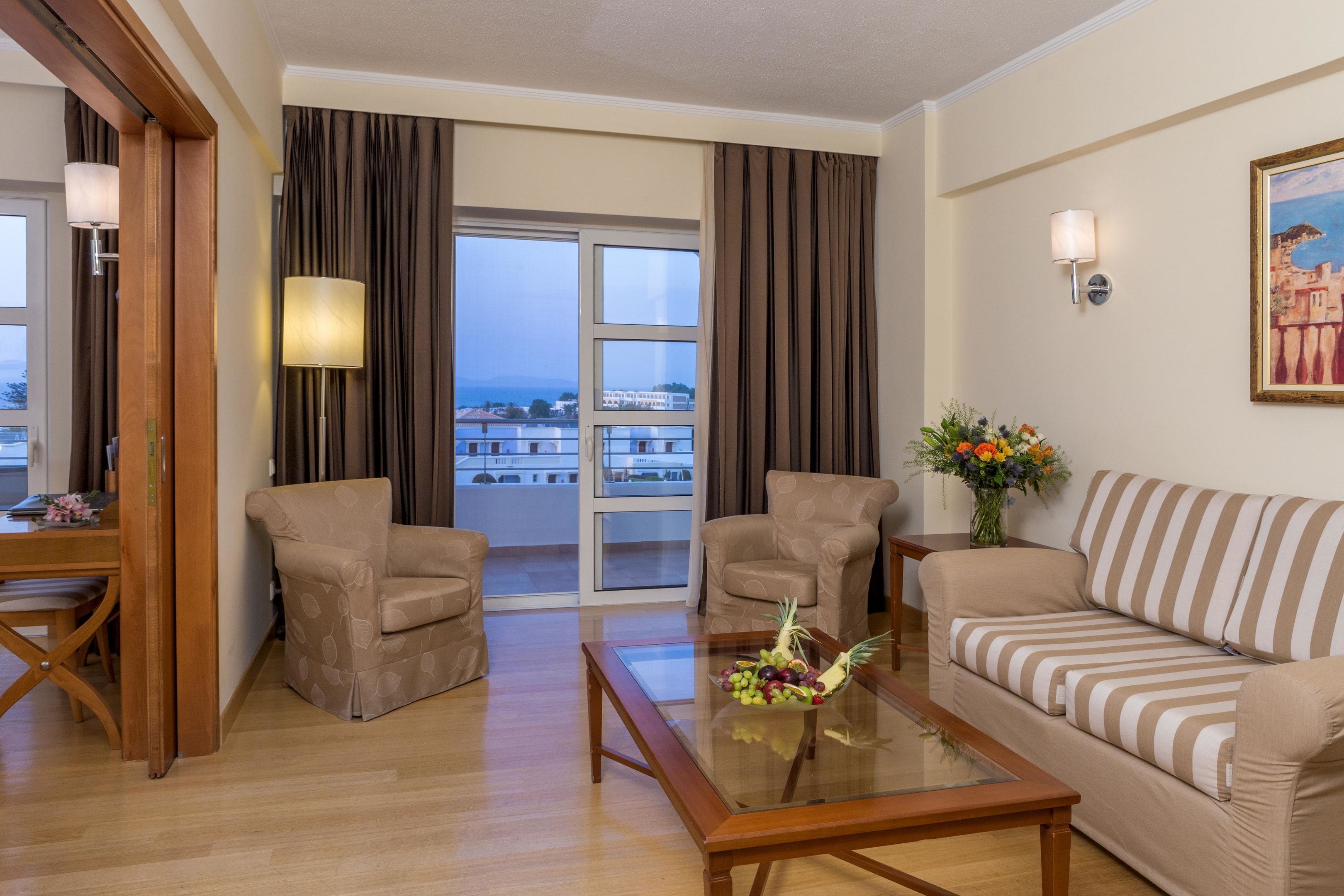 A hotel room with a sofa, two armchairs, a dining table, a glass coffee table with fruit, a vase with flowers, a painting on the wall, and a view of the sea through large windows.
