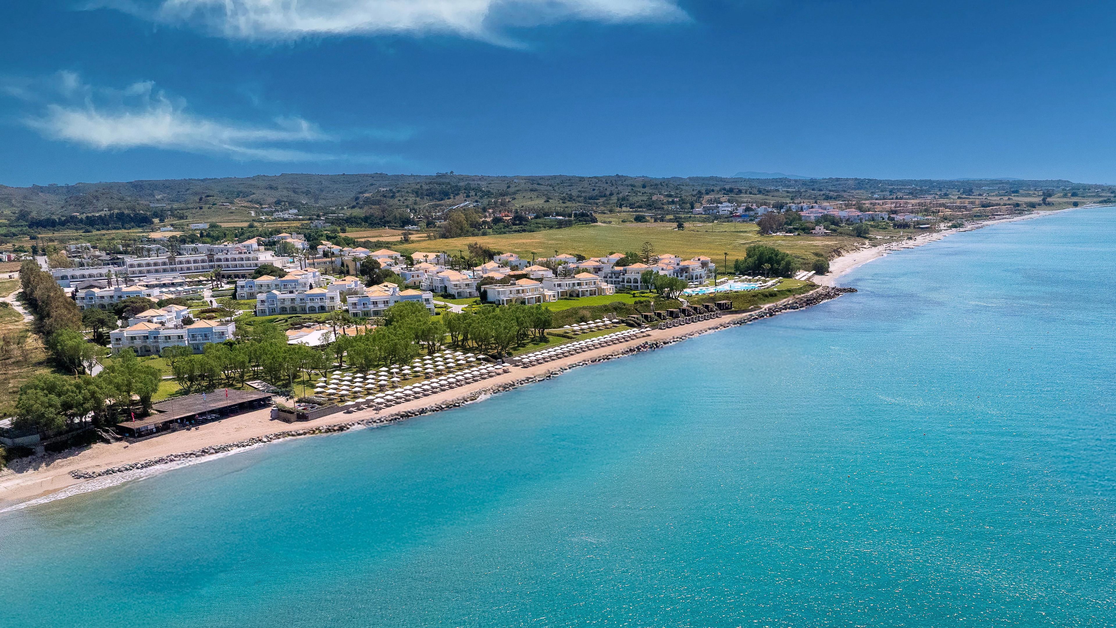 Un resort costero con villas frente a la playa, una playa de arena y un mar azul cristalino.