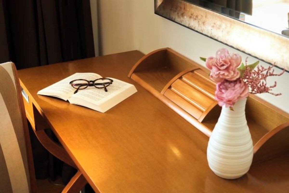 Una mesa de madera con un jarrón blanco lleno de flores rosas, un par de gafas y un documento.