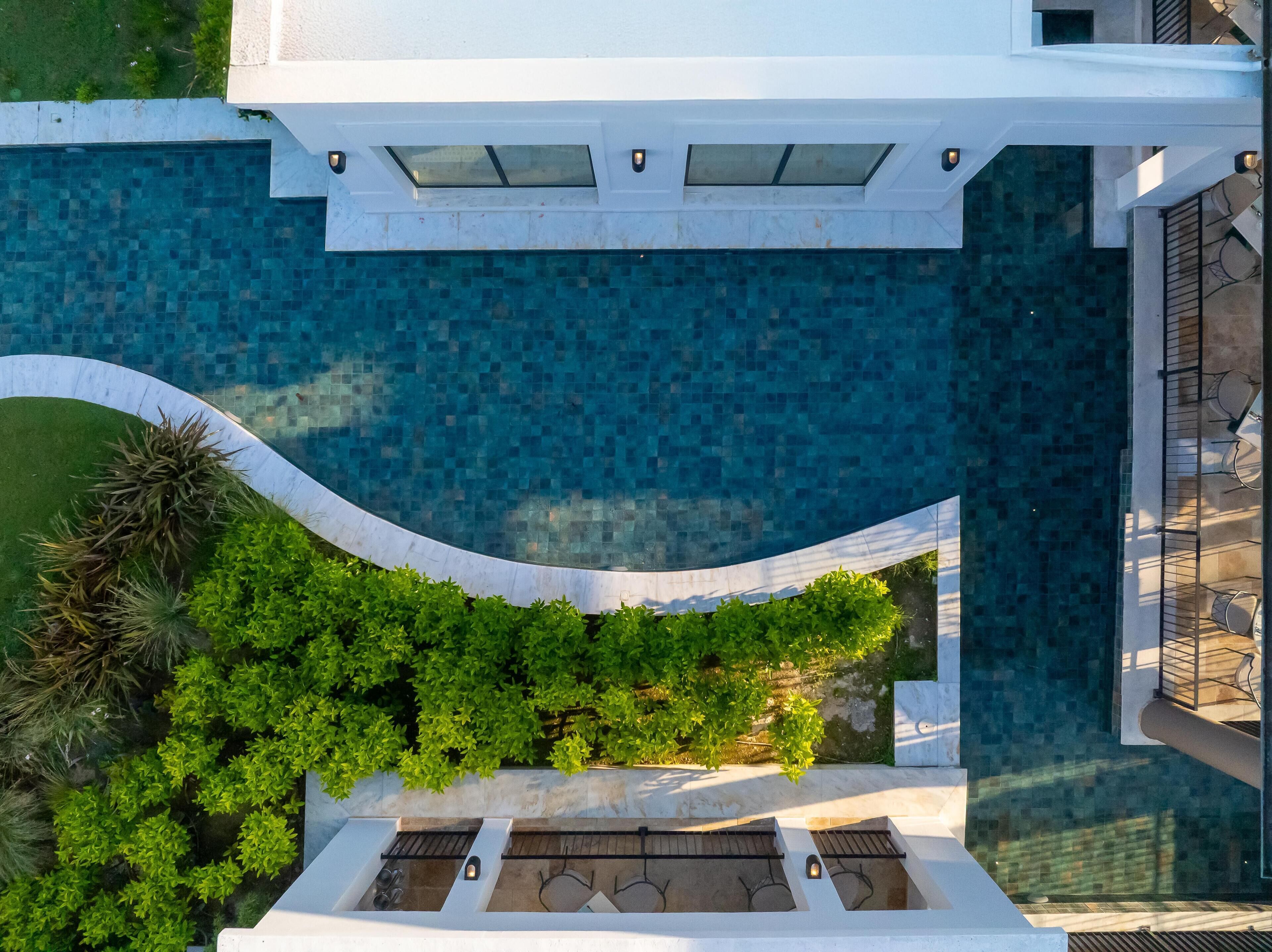 Vista aerea di una piscina a forma curva, circondata da vegetazione rigogliosa e un edificio bianco con tetto in tegole.