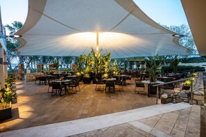 Ein Terrassenbereich im Freien mit Tischen, Stühlen, einem großen Sonnensegel und Topfpflanzen.