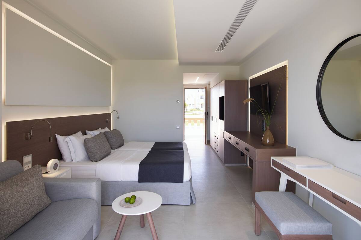 A modern hotel room with a bed, a sofa, a small table with fruit, a vanity with a mirror, and a desk.