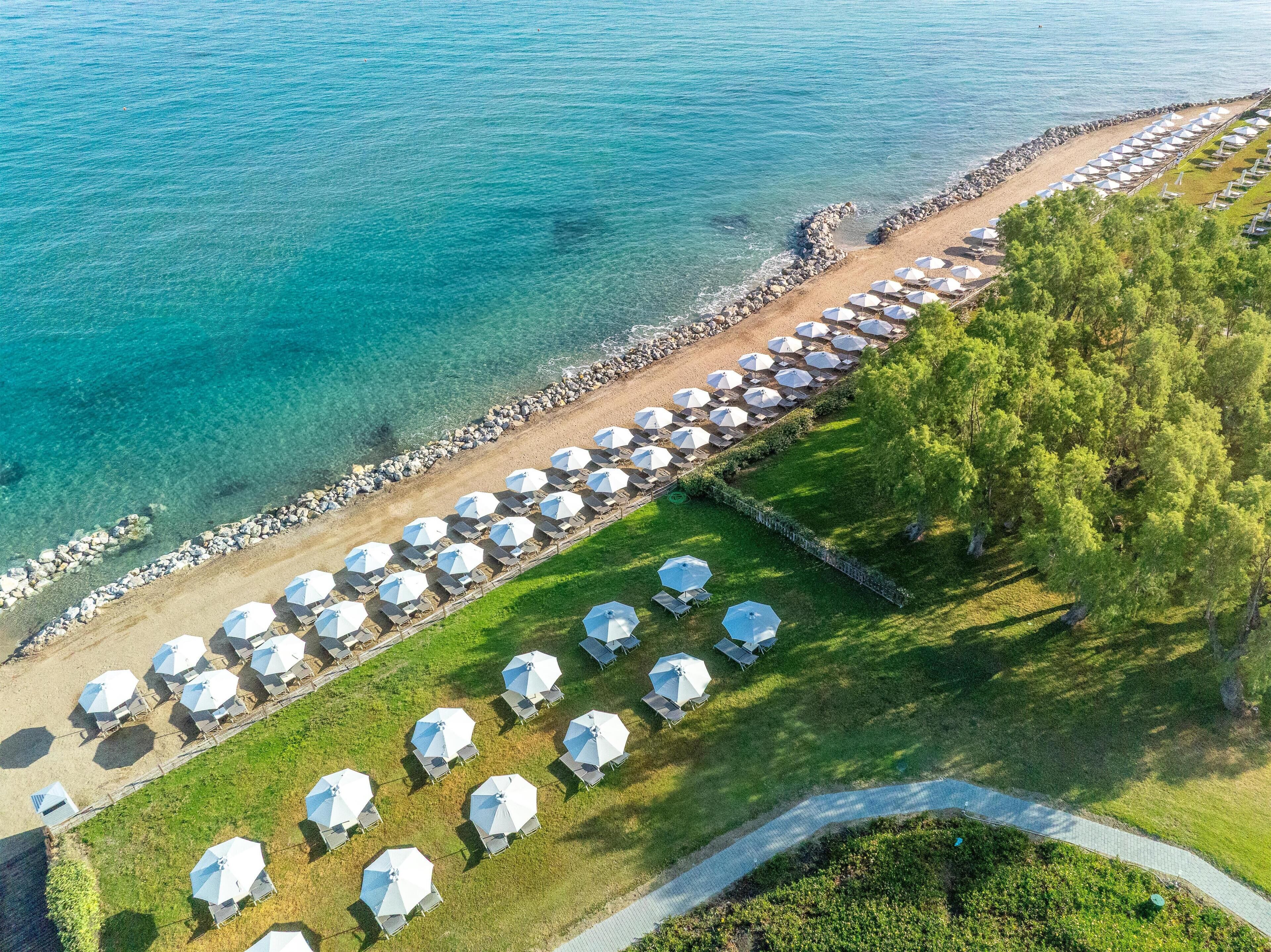 An aerial view of a beachside resort with rows of umbrellas and lounge chairs along the shoreline, a paved walkway, and a rocky breakwater.