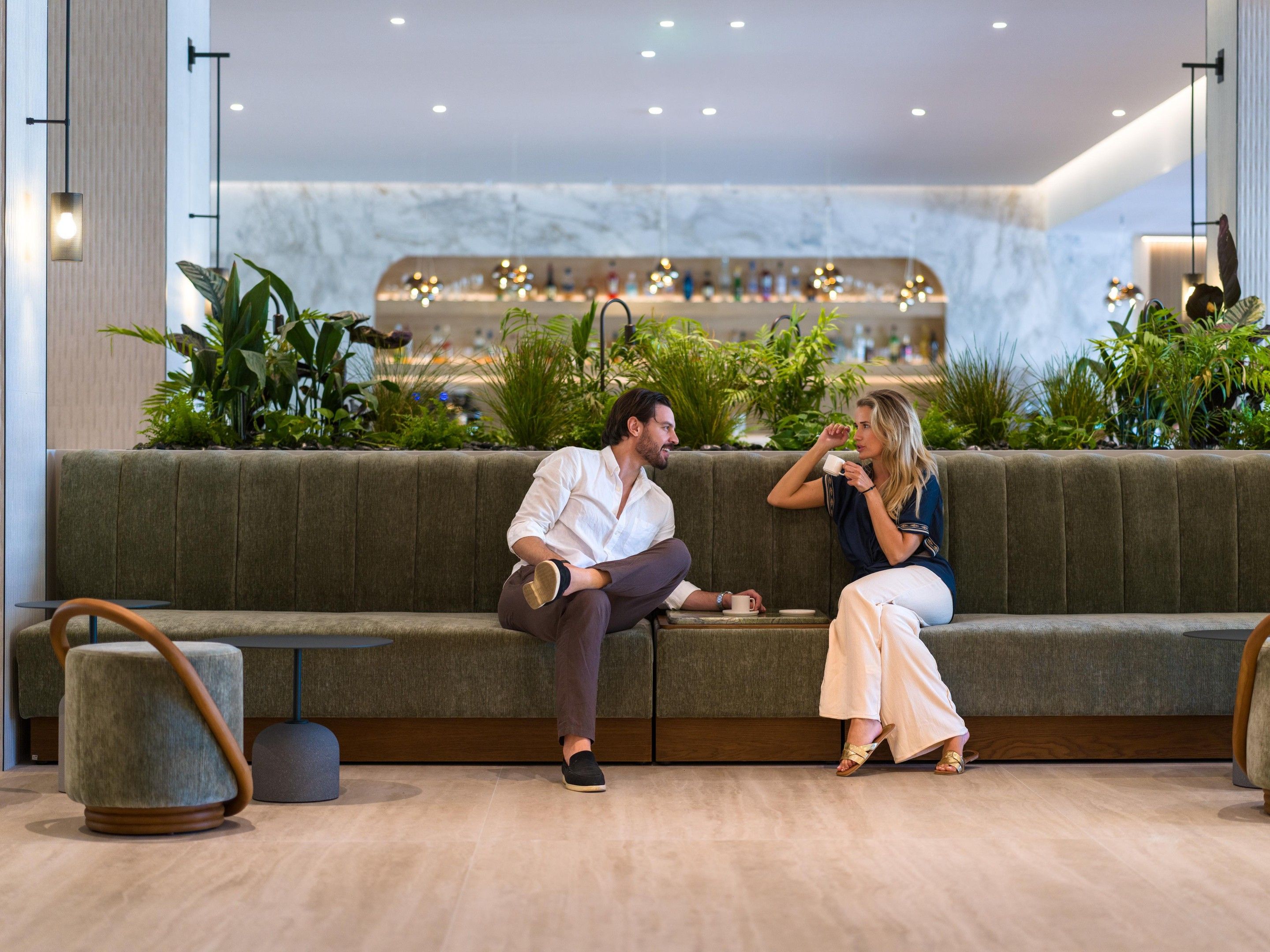A modern lounge area with a sofa, a small table, and a seating area with a view of a bar and plants.