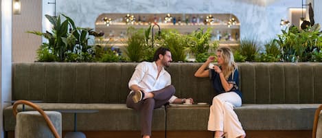 A modern lounge area with a sofa, a small table, and a seating area with a view of a bar and plants.