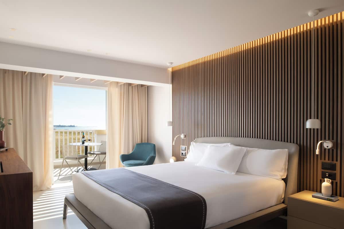 A modern hotel room with a large bed, a desk, a chair, and a balcony with a view.
