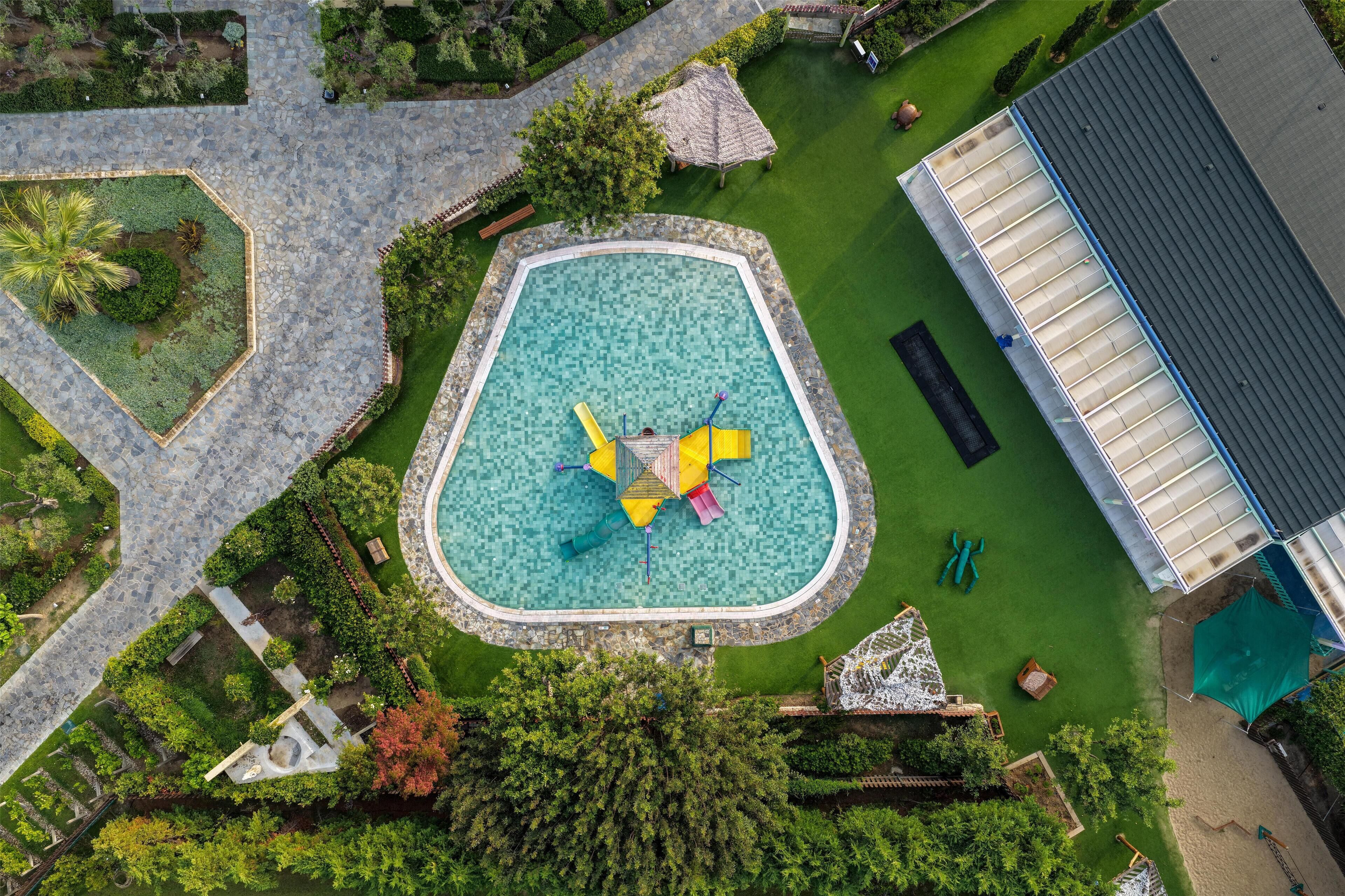 An aerial view of a swimming pool surrounded by a landscaped garden, with a covered structure to the right.