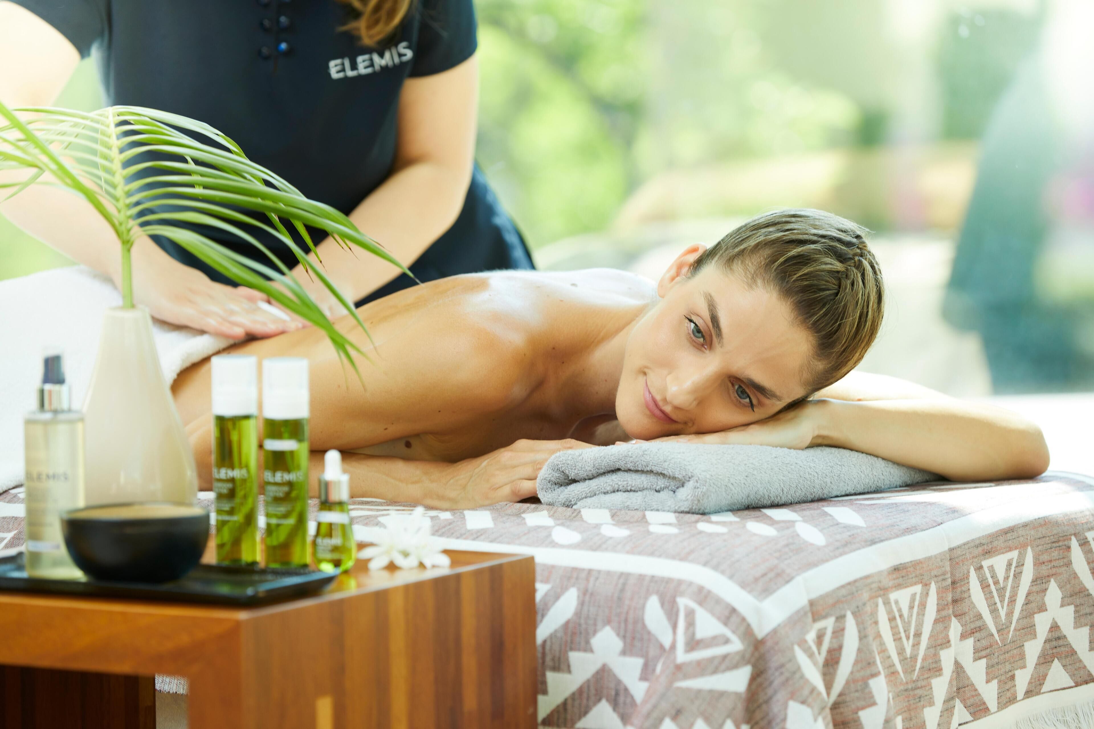 A massage therapy session with a client lying on a patterned bed, a therapist in a black shirt, and various spa products on a wooden table.