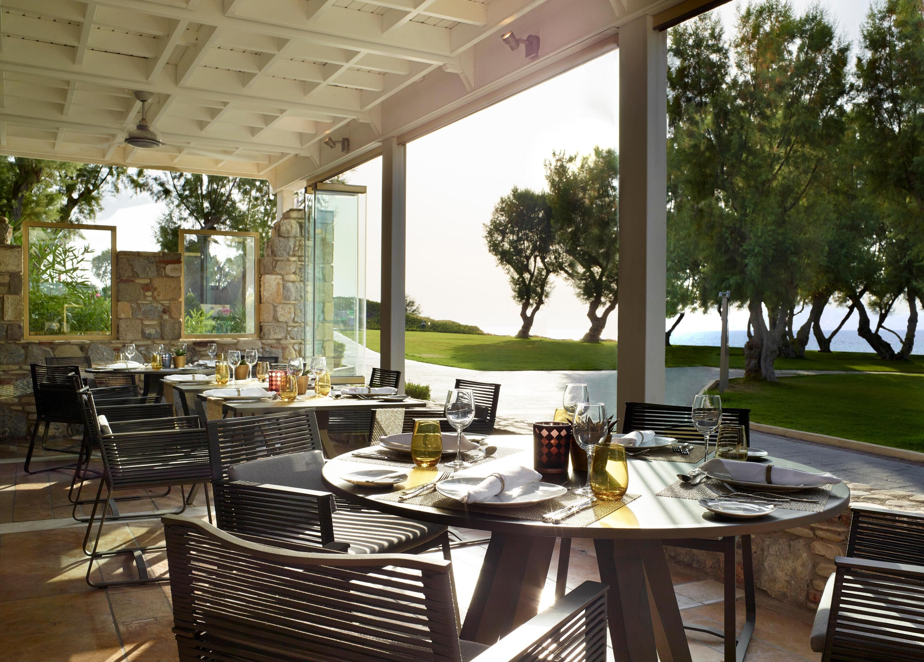 An outdoor dining area with tables and chairs, a stone wall, and a view of trees and a body of water.